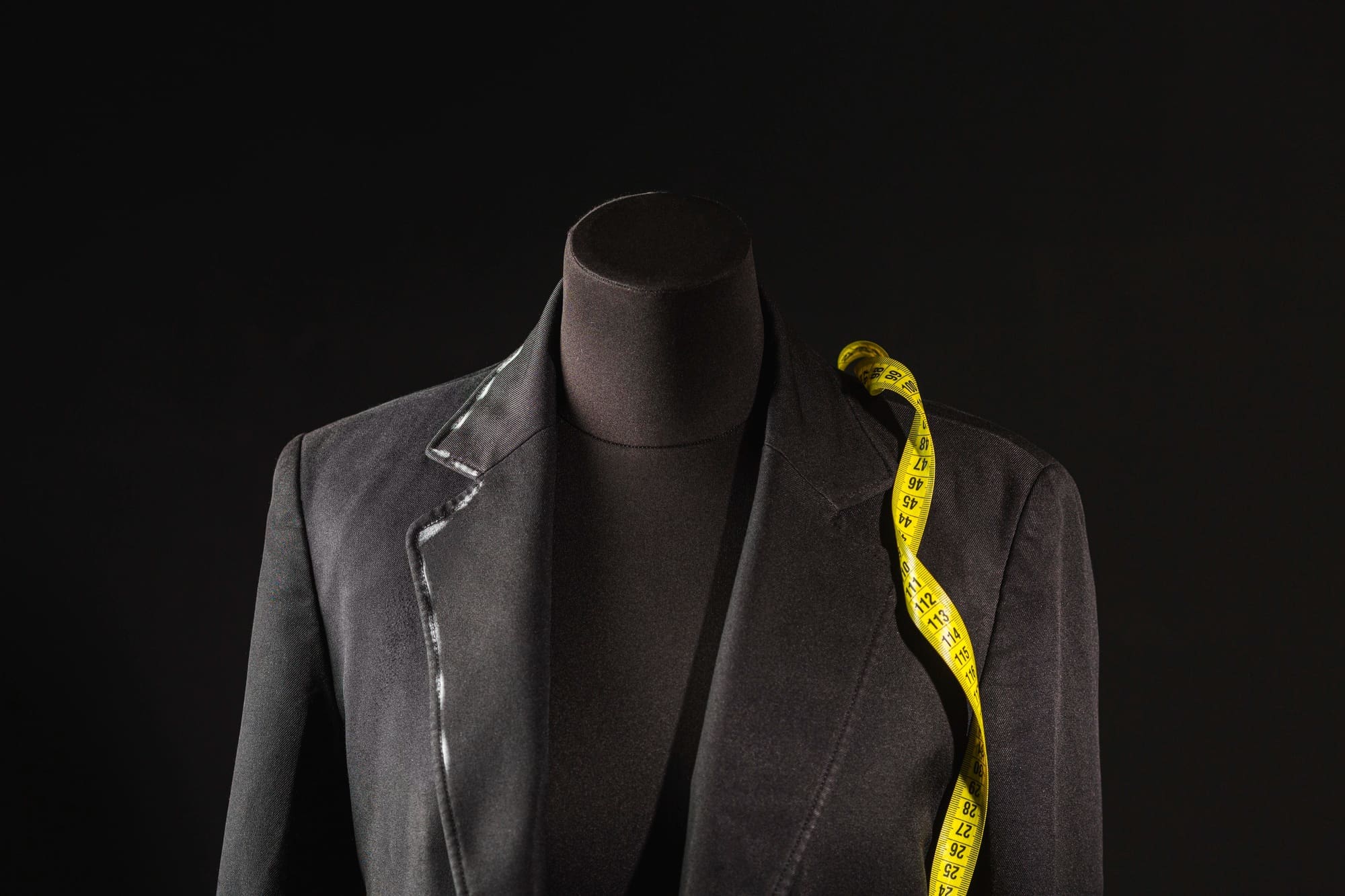 A black tailor&rsquo;s mannequin dressed in a dark blazer with a yellow measuring tape draped over one shoulder, set against a black background&mdash;perfect for showcasing expert leather garment cleaning Syracuse services.