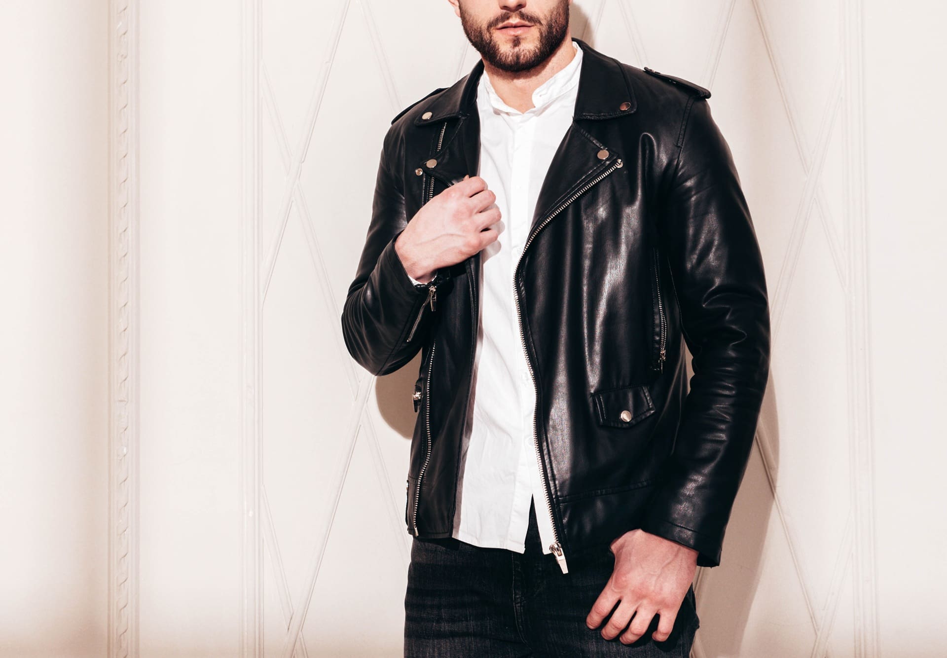 A man in a black leather jacket over a white shirt stands against a beige wall, with his left hand touching the jacket. His face is partially out of frame, much like newly cleaned gowns at Wedding dress cleaning Manlius&mdash;spotless and sharp.