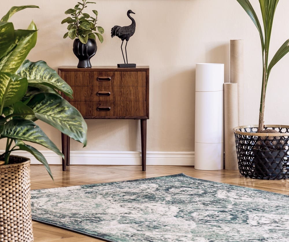 A mid-century wooden cabinet with plants and a crane sculpture stands beside a green patterned rug in a bright, modern room—an inviting spot inspired by the timeless style of alterations and tailoring Syracuse.